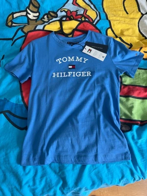 Blå t-shirt från Tommy Hilfiger - Snygg blå t-shirt från Tommy Hilfiger med klassisk logga broderad på bröstet. T-shirten har rund hals och korta ärmar. Tillverkad i mjuk bomull för skön känsla och stilren look.