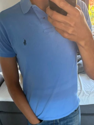 Blå pikétröja från Polo Ralph Lauren - Snygg ljusblå pikétröja från Polo Ralph Lauren med klassisk krage och korta ärmar. Tröjan har den ikoniska loggan broderad på bröstet och är tillverkad i mjuk bomull som känns skön mot huden. Perfekt till jeans eller shorts för en clean look.