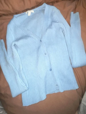 Ljusblå ribbad kofta från H&M - Snygg ljusblå ribbad kofta från H&M med v-ringning och knäppning framtill. Långärmad modell i mjukt, stretchigt material som sitter skönt på kroppen. Perfekt att slänga över en topp eller bära som den är.