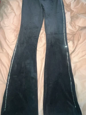 Svarta utsvängda leggings med strass - Snygga svarta leggings i mjukt sammetstyg med utsvängda ben. Längs sidorna finns en rad med glittrande strass-stenar som ger extra bling. Perfekta för dig som vill sticka ut lite extra och gillar glam.