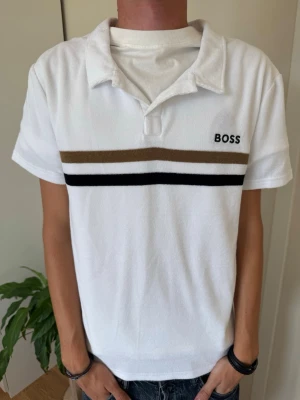 Vit kortärmad skjorta från BOSS - Vit skjorta från BOSS med korta ärmar och klassisk krage. Två horisontella ränder i brunt och svart över bröstet samt BOSS-logga broderad på vänster sida. Perfekt för en clean och stilren look.
