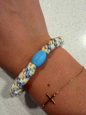 Kknekki hårband - Säljer ett flätat armband i pastellfärger med en blå oval detalj där det står 'knekki Armbandet har inslag av ljusgul, beige och ljusblå och är gjort av syntetmaterial med en glansig finish. Perfekt för dig som gillar färgglada accessoarer.