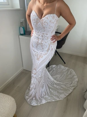 Vit långklänning  - Elegant vit långklänning med tunna axelband med en sweetheart neckline. Klänningen har ett vackert spetsmönster över hela tyget och en lång släp baktill, full med paljetter. Perfekt för förlovningen, bröllop eller bal! Klänningen är ifrån Tina Holly (Australien). Använd en gång! Nypris 3000