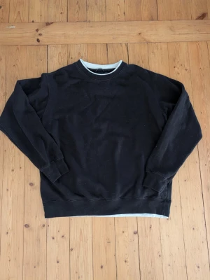 Svart sweatshirt med rund hals - En basic svart sweatshirt med rund hals och långa ärmar. Tröjan har ribbade muddar vid ärmslut och nederkant samt en vit detalj i halsringningen. Perfekt för en avslappnad och stilren look.