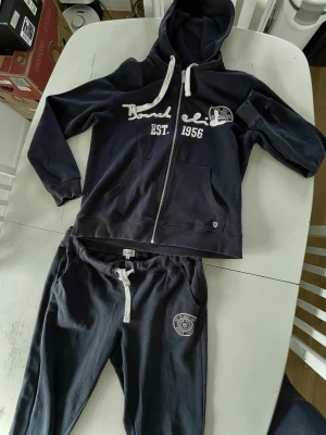 Mörkblå hoodie med dragkedja och tryck - Säljer en mörkblå hoodie med vit dragkedja, huva och vita snören. Tryck på bröstet med texten 'Board Club EST. 1956' och ett märke på vänster sida. Hoodien har två fickor framtill och ribbade muddar. Perfekt för chill dagar eller träning.