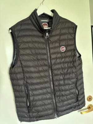 Svart dunväst från Colmar storlek S - Snygg svart dunväst från Colmar med quiltad design och hög krage. Västen har två dragkedjeförsedda fickor och en tydlig Colmar-logga på bröstet. Perfekt att slänga över en hoodie eller tröja för extra värme.