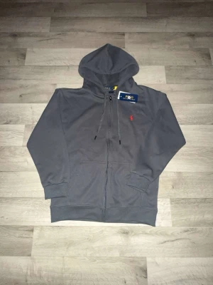 Grå hoodie från Polo Ralph Lauren - Snygg grå hoodie från Polo Ralph Lauren med dragkedja framtill och klassisk röd logga broderad på bröstet. Hoodien har justerbar huva med snören och två fickor framtill. Perfekt för en chill och stilren look.