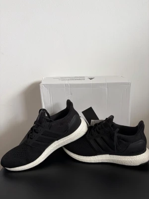 Svarta Adidas Ultraboost sneakers - Snygga svarta Adidas Ultraboost sneakers med stickad ovandel och vit, responsiv sula. Skorna har klassiska tre ränder på sidan och svart hälkappa. Perfekta för dig som gillar stilrena och bekväma sneakers med modern design.