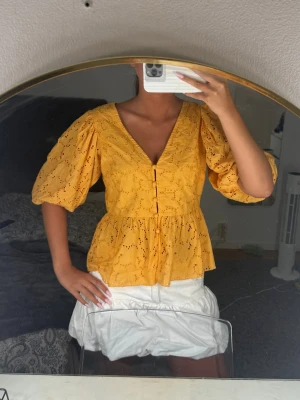 blus med broderade detaljer - Superfin  blus med v-ringning, puffärmar och broderade håldetaljer. Blusen har knappar framtill och en lätt volang nertill som ger en söt siluett. Perfekt för dig som vill ha en färgglad och trendig topp till sommaren.