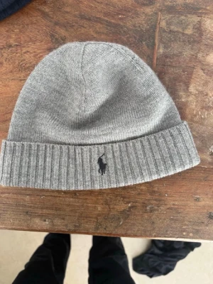 Grå ribbstickad mössa från Polo Ralph Lauren - Snygg grå mössa från Polo Ralph Lauren med ribbstickad kant och klassisk broderad logga framtill. Tillverkad i mjukt stickat material som håller dig varm och stilren under kalla dagar. Perfekt accessoar för en clean och avslappnad look.