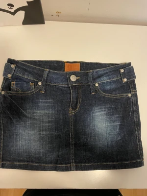 Mörkblå jeanskjol med fickor - Säljer en snygg mörkblå jeanskjol i kort modell med slitningar och kontrastsömmar. Kjolen har klassiska fem fickor, bälteshällor och knappstängning fram. Perfekt till en avslappnad och trendig look.