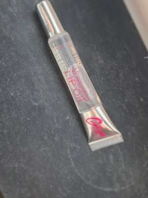 Lip oil glam of Sweden  - Fuktgivande läppolja i genomskinlig tub med silvrig kork och rosa text. Oljan är transparent och ger en glansig finish. Perfekt för att återfukta och ge läpparna en snygg glansig look.