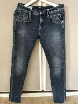 Replay blå slim fit jeans - Snygga blå slim fit jeans från Replay med klassisk femficksdesign och diskreta slitningar. Jeansen har en något tvättad look och smal passform som sitter tajt längs benen. Perfekta för en trendig och avslappnad stil.