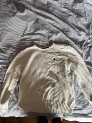 Vit stickad tröja från Polo Ralph Lauren - Klassisk vit stickad tröja från Polo Ralph Lauren i 100% bomull. Tröjan har rund halsringning, ribbstickade muddar och den ikoniska lilla loggan broderad på bröstet. Perfekt för en clean och stilren look.