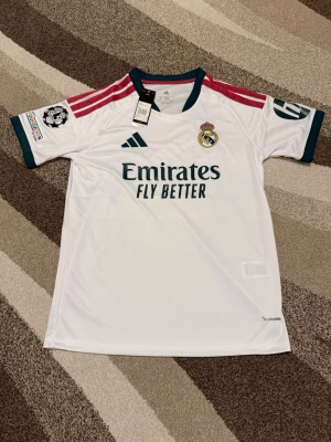 Real Madrid 26-27 hemmatröja - Real Madrid 26-27 hemmatröja   Storlek: M Namn och nummer: Ingen Badge: Champions League  Beskrivning: Tröjan är helt ny och felfri.  Säljs av ICEKIT icekit.se