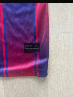 Randig Dri-FIT tröja från Nike - Säljer en sportig Dri-FIT tröja från Nike med vertikala ränder i lila, vinrött och blått. Tröjan har en lätt och funktionell känsla, perfekt för träning eller match. Materialet är snabbtorkande och andas bra.
