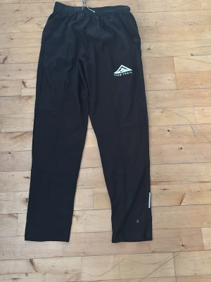 Svarta Nike Trail träningsbyxor - Säljer ett par svarta träningsbyxor från Nike Trail med Dri-FIT ADV-material som transporterar bort fukt. Byxorna har resår i midjan, tryckt logga på låret och knappar vid bensluten för justering. Perfekta för löpning eller utomhusträning.