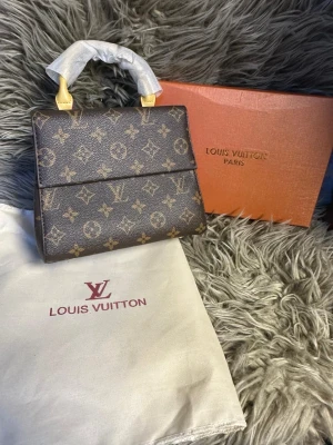  monogram handväska - Snygg handväska från i klassiskt brunt monogrammönster. Väskan har guldfärgade detaljer, topphandtag och är tillverkad i canvas med läderdetaljer. Kommer med dustbag och originalkartong. Perfekt accessoar för dig som gillar lyxig och tidlös design.