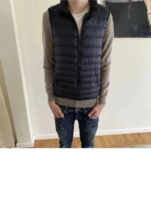 Mörkblå lättviktsdunväst - Uniqlo väst i storlek S. Snygg mörkblå dunväst med quiltad design och dragkedja framtill. Västen har hög krage och är tillverkad i ett lätt material som ger bra värme utan att kännas klumpig. Perfekt att slänga över en hoodie eller tröja för extra lager.
