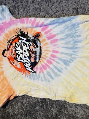 Färgglad tie dye t-shirt med tryck - Säljer en riktigt färgglad tie dye t-shirt med ett stort tryck av en tecknad kille på sparkcykel och texten 'ISAK HUDDEN' på bröstet. T-shirten har korta ärmar och är gjord i mjuk bomull. Perfekt för dig som gillar streetstyle och unika plagg!