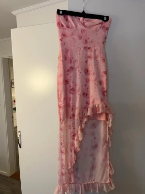 Rosa blommig långklänning med volang - Söt långklänning i ljusrosa med blommigt mönster och volangdetaljer längs nederkanten och sidan. Klänningen är axelbandslös och har en asymmetrisk skärning framtill. Perfekt för sommarens alla tillfällen.