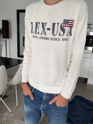 Vit stickad tröja med tryck - Vit stickad tröja med rund hals och långa ärmar. På bröstet finns ett stort tryck med texten 'LEX-USA' och en liten amerikansk flagga. Tröjan har ribbade muddar vid ärmslut och nederkant. Perfekt för en avslappnad stil. Tröjan passar en vanlig M