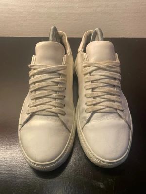Vita låga sneakers i läder - Säljer ett par klassiska vita sneakers i läder med matchande vita skosnören och diskret design. Skorna har en rund tå och platt sula, perfekta för en clean och stilren look. Passar till det mesta och är enkla att styla.