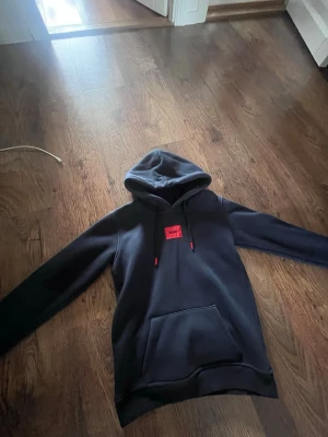 Mörkblå Hugo boss hoodie med röd logga - Snygg mörkblå hoodie med huva och stor magficka. På bröstet sitter en röd patch med texten 'HUGO'. Hoodien har dragsko i huvan och ribbade muddar vid ärmslut och nederkant. Perfekt för en avslappnad och trendig look.