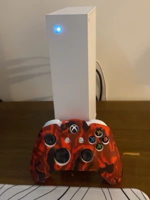 Vit Xbox Series S med röd kontroll - Säljer en vit Xbox Series S med en unik handkontroll i röd och svart camo. Konsolen har en stilren, rektangulär design och flera anslutningsmöjligheter på baksidan. Perfekt för gaming och streaming.