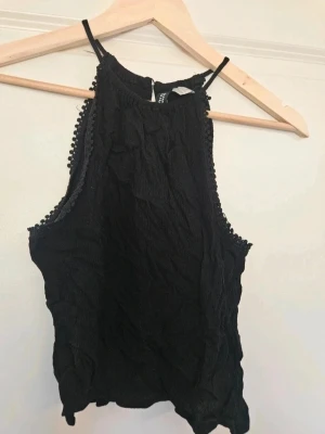Svart ärmlös linnetopp H&M 32 - Säljer en svart ärmlös linnetopp från H&M med smala axelband och dekorativa små bollar längs kanterna. Toppen har en lätt rynkad struktur och hög halsringning. Perfekt att styla med jeans eller kjol. E4