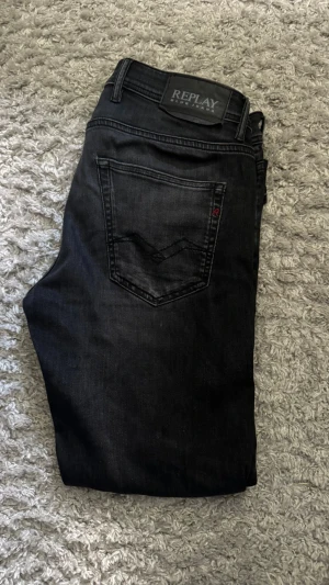 Svarta Replay jeans med smal passform - Snygga svarta jeans från Replay med rak passform och klassisk femficksdesign. Jeansen har diskreta slitningar och en svart läderpatch med Replay-logga bak i midjan. Tillverkade i mjukt denimtyg som ger en bekväm känsla och stilren look. Använt fåtal gånger så nästan nyskick