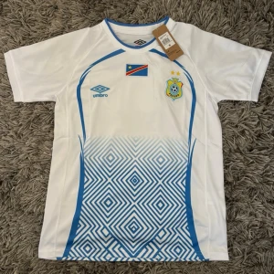 Vit Umbro fotbollströja DR Kongo - Säljer en vit fotbollströja från Umbro med DR Kongos landslagsemblem och flagga på bröstet. Tröjan har blå detaljer och ett geometriskt mönster nedtill. Perfekt för fotboll eller träning!