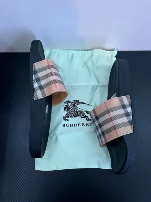 Burberry tofflor  - Snygga sandaler från Burberry med klassiskt beige, svart och rött rutigt band över foten. Sulan är svart och har Burberry-logga på sidan. Perfekta för sommaren och ger en lyxig touch till din outfit. Kommer med originalpåse.