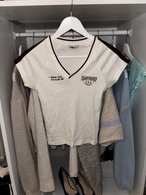 Vit v-ringad t-shirt med tryck - Vit t-shirt med v-ringning och vinröda detaljer på axlar och krage. Tryck på bröstet med texten 'CHAMPIONSHIP' och 'SOUTH CLUB'. Den är lite liten i storleken.