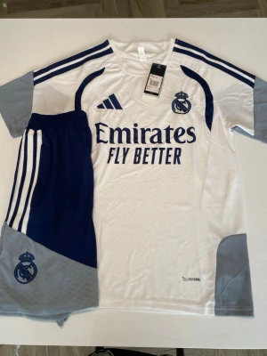 Real Madrid matchställ Adidas S - Säljer ett Real Madrid matchställ från Adidas i storlek S. Setet består av en vit och blå tröja med klubbmärke, sponsortryck och Climacool-material samt matchande shorts i blått och grått med klubbmärke och klassiska Adidas-ränder.
