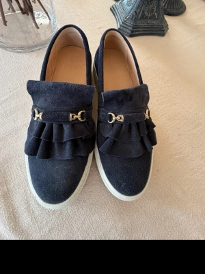 Mörkblå loafers med volang och gulddetalj - Snygga mörkblå loafers i mocka med volang framtill och guldfärgad metallspänne. Skorna har vit platåsula och en stilren design som sticker ut. Perfekt för dig som vill ha något unikt och trendigt i garderoben. Nypris ca 2000