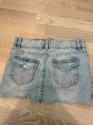 Ljusblå jeanskjol med rå kant - Säljer en kort ljusblå jeanskjol med rå, fransig kant och slitningar. Kjolen har två bakfickor med rosa broderi, bälteshällor och knappstängning fram. Perfekt för dig som gillar en avslappnad och trendig stil.