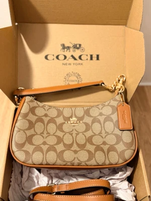 Coach brun teri shoulder bag signature canvas - Snygg brun handväska från Coach med klassiskt monogrammönster och detaljer i ljusbrunt läder. Väskan har guldfärgade metalldetaljer, avtagbar axelrem och Coach-tag. Perfekt storlek för det viktigaste och en stilren look.