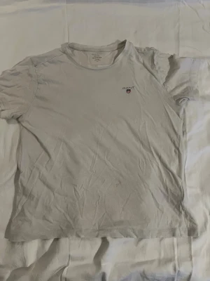 Vit t-shirt från GANT med logga - Klassisk vit t-shirt från GANT med liten broderad logga på bröstet. T-shirten har rund halsringning och korta ärmar. Tillverkad i mjuk bomull för en skön känsla. Perfekt basplagg till garderoben.