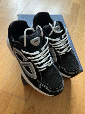 Svarta sneakers med vita detaljer - Snygga svarta sneakers med vita och grå detaljer samt vita skosnören. Skorna har en sportig design med mesh och läderpaneler, samt en tjock vit sula för extra komfort. Perfekta för dig som vill ha en exklusiv och trendig look.