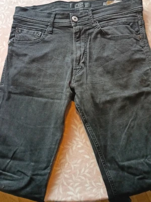Svarta raka jeans med fem fickor, det är Replay jeans - Säljer ett par svarta jeans med klassisk femficksmodell och raka ben. Jeansen har knappgylf och bälteshällor. Materialet är denim med en mjuk känsla och lätt tvättad look. Perfekta för dig som gillar en enkel och stilren stil.