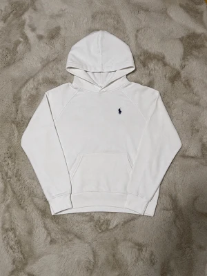 Vit hoodie från Polo Ralph Lauren - Snygg vit hoodie från Polo Ralph Lauren med klassisk marinblå logga broderad på bröstet. Hoodien har huva, känguruficka och ribbade muddar vid ärmar och nederkant. Tillverkad i mjukt bomullsmaterial som är skönt mot huden.