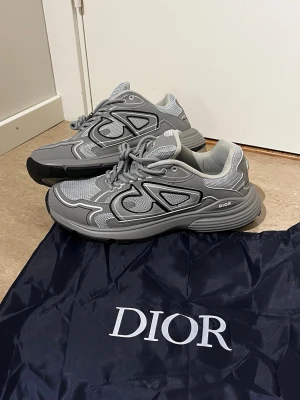 Grå Dioor sneakers med mesh och detaljer - Snygga grå sneakers från Dioor med coola svarta och vita detaljer. Skorna har en chunky sula, meshpaneler och syntetiska överdrag för en modern look. Perfekta för dig som gillar streetwear och exklusiva märken.