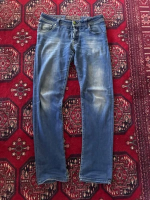 Blå jeans från Jacob Cohën - Snygga blå jeans från Jacob Cohën med klassisk femficksmodell och diskret slitning på låren och knäna. Jeansen har raka ben och detaljerade sömmar. Tillverkade i mjukt denimtyg för en bekväm känsla. Jeansen är använda en hel del, och har små hål samt reparationer. Och såklart ett större hål på insidsn av fickan, vilket alltså inte syns eller påverkar fickans funktion. Skick: 7,25/10.