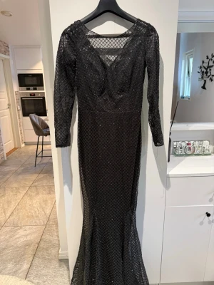 Svart långklänning med glitter - Snygg svart långklänning med glittrigt rutmönster och långärmad design. Klänningen har en v-ringad urringning och meshdetaljer upptill. Perfekt för dig som vill sticka ut på festen med en elegant och glamorös look.