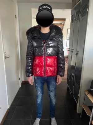 Mörkblå/röd pufferjacka Tommy Hilfiger - Snygg pufferjacka från Tommy Hilfiger i glansigt material med mörkblå överdel och röd nederdel. Jackan har huva, dragkedja framtill och en liten logga på ärmen. Perfekt för kalla vinterdagar och riktigt trendig look.