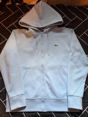 Ljusblå zip hoodie från Lacoste - Snygg ljusblå hoodie från Lacoste med dragkedja framtill och klassisk krokodil-logga på bröstet. Tröjan har huva med dragsko, två fickor fram och ribbade muddar vid ärmar och nederkant. Perfekt för en chill och stilren look.