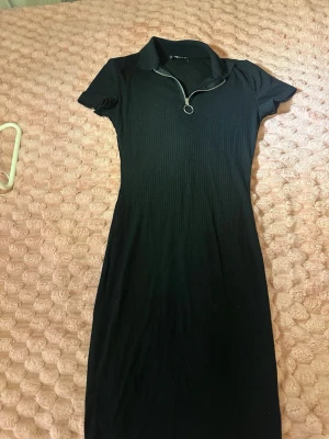 Svart ribbad klänning från Sister XS - Svart ribbad klänning från Sister i storlek XS. Klänningen har korta ärmar, krage och dragkedja framtill med ringdetalj. Materialet är stretchigt och ger en slim passform. Perfekt för dig som gillar en enkel men snygg stil.