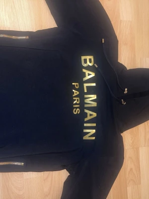  Balmain hoodie - Snygg svart hoodie från Balmain Paris med stort guldfärgat tryck på bröstet och guldfärgade dragkedjor vid sidorna. Klassisk huva med dragsko och metalländar. Perfekt för dig som vill sticka ut med exklusiv streetstyle.