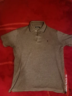 Grå pikétröja från Polo Ralph Lauren - Klassisk grå pikétröja från Polo Ralph Lauren med krage, knappar och diskret logga på bröstet. Tröjan har korta ärmar och är tillverkad i ett mjukt bomullsmaterial. Perfekt för en avslappnad och stilren look.